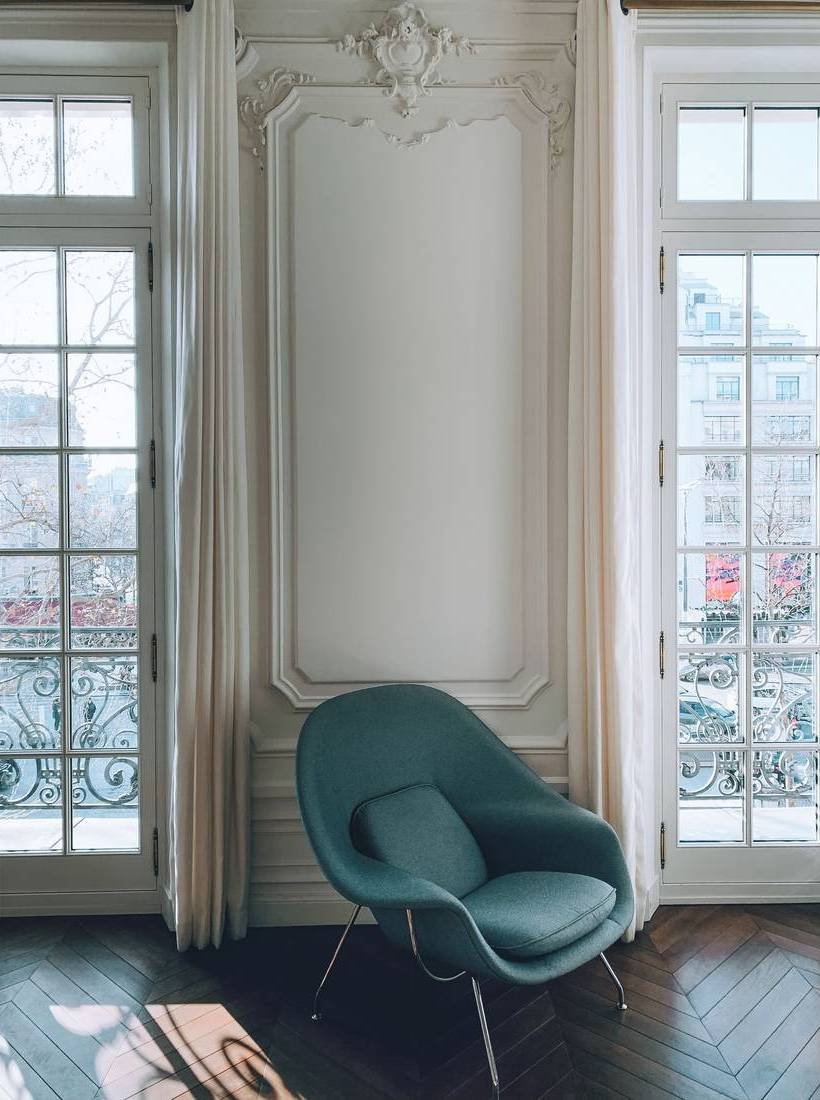 A moulded Parisian-style salon wall between French windows, with a teal lounge chair on a herringbone floor